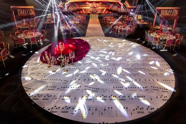 Circular dance floor printed with musical notation, lit by moving lights, with red-dressed tables beyond, MY Event Design