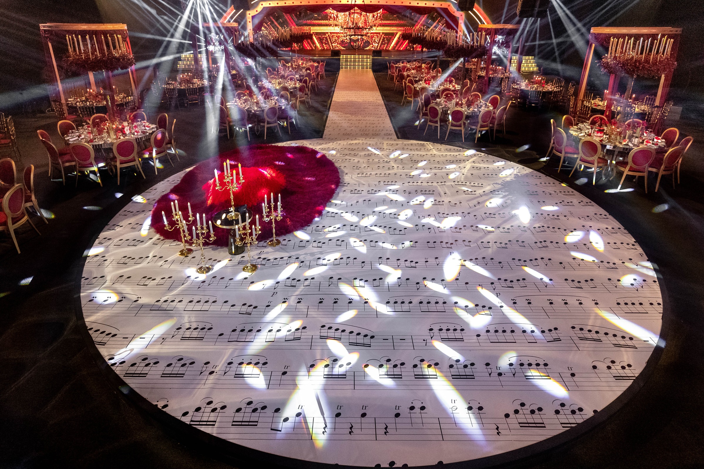 Circular dance floor printed with musical notation, lit by moving lights, with red-dressed tables beyond, MY Event Design
