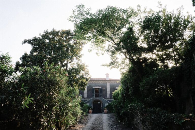 The venue’s stone entrance framed through an arch of trees, shuttered windows catching the last evening light, My Sicilian Wedding