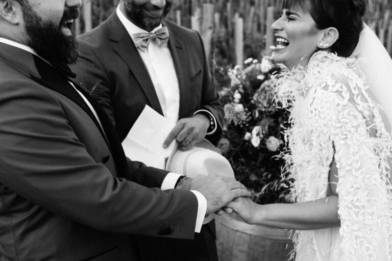 The groom shares a laugh mid-vows, the bride laughing in an ostrich-feather gown, a vineyard row behind them, My Sicilian Wedding