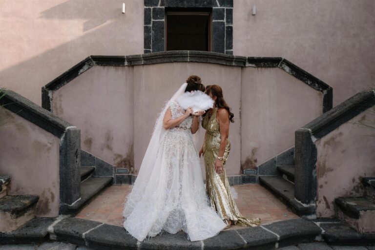The bride and her mother share a private moment behind a garden wall, a white feather fan raised between them, My Sicilian Wedding
