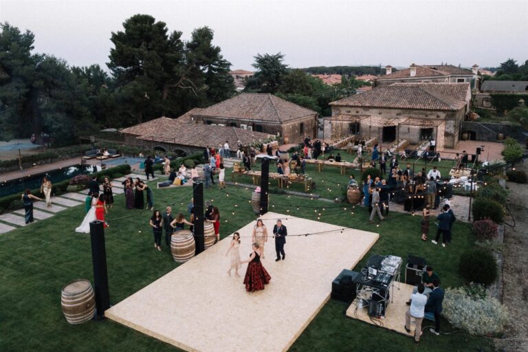 Aerial view of the reception in full swing: a lit dance floor, a pool, and guests scattered across the lawn beneath string lights, My Sicilian Wedding
