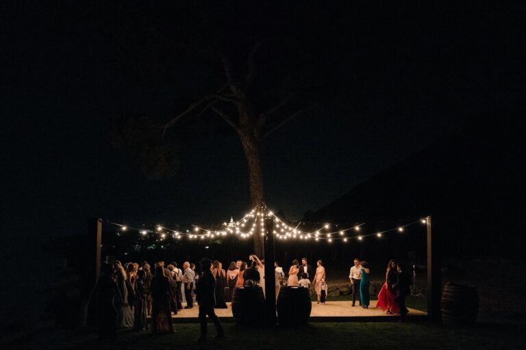 Guests dance beneath a canopy of string lights strung from an old tree, the hillside and town lights visible in the dark beyond, My Sicilian Wedding
