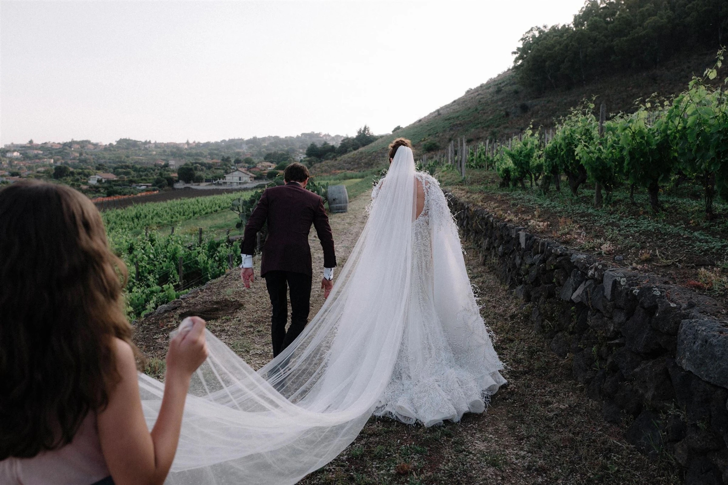 A bride and groom walk hand in hand along a dry-stone wall between rows of Etna vineyard, her cathedral veil carried behind her by a bridesmaid, My Sicilian Wedding
