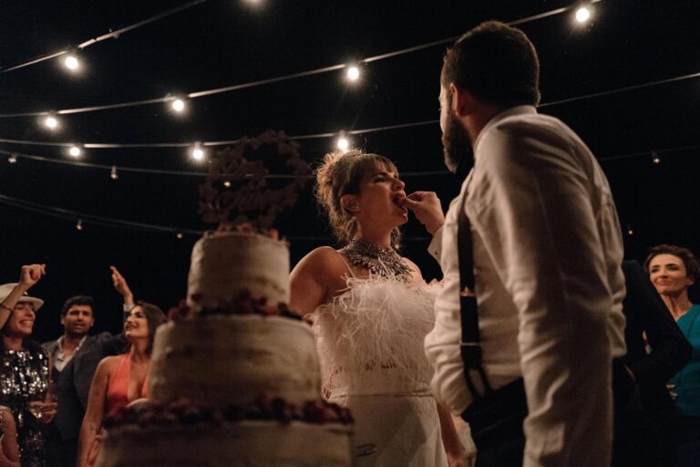 The groom feeds the bride a bite of their tiered wedding cake beneath strings of festoon lighting as guests cheer, My Sicilian Wedding