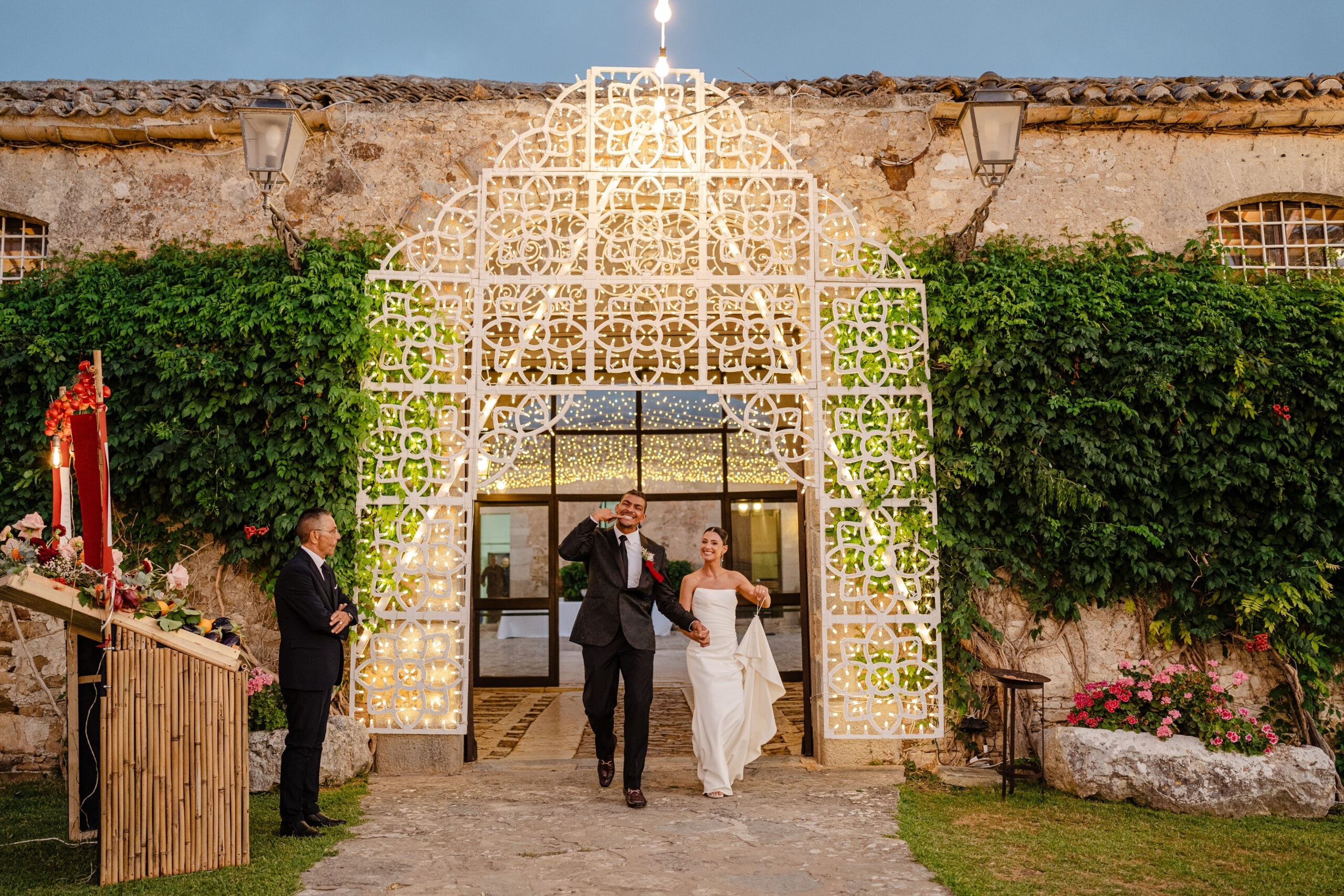 Walking out through a lit luminaria arch set into an ivy-covered wall, My Sicily Wedding