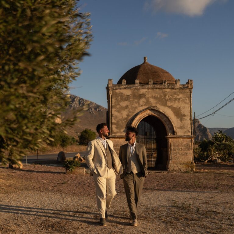 Two grooms walking from a small domed chapel, olive branches in the foreground, My Sicily Wedding