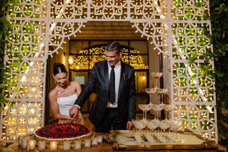 Cutting the tart in front of the luminaria, a tower of coupes alongside, My Sicily Wedding