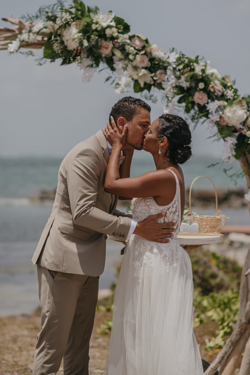 A couple kissing in front of a floral arch on a sandy beach, Myl'Evasions