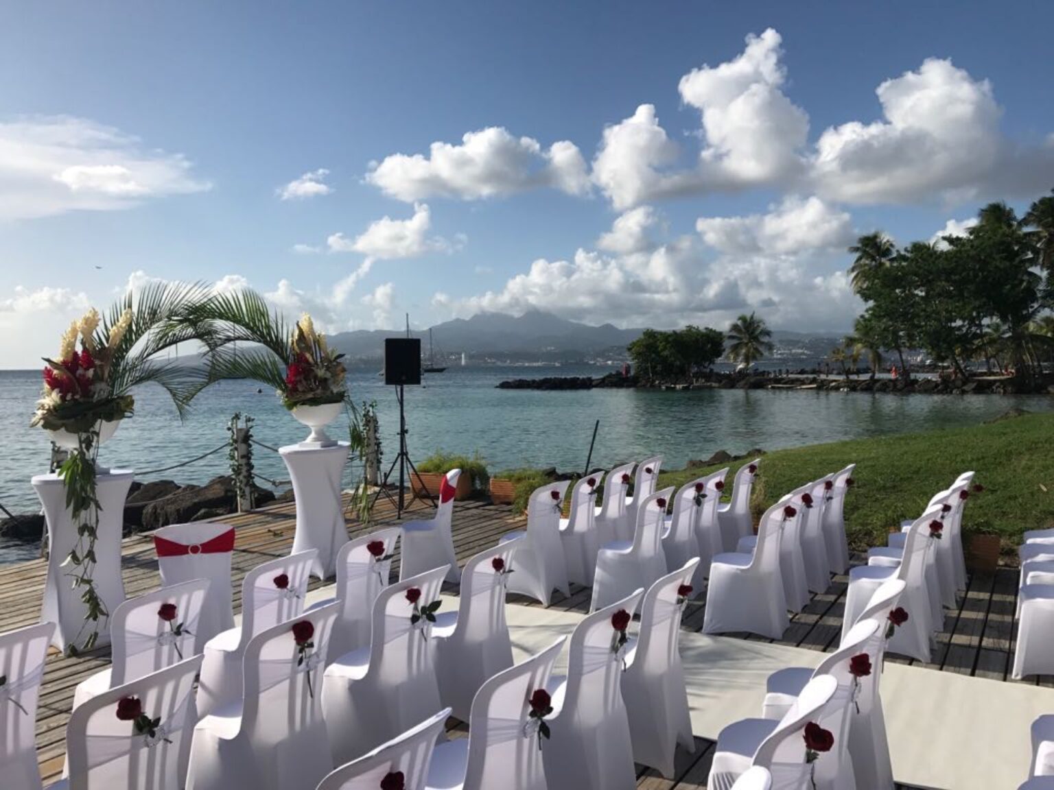Rows of white covered chairs facing a floral arch on a seafront terrace overlooking a bay, Myl'Evasions
