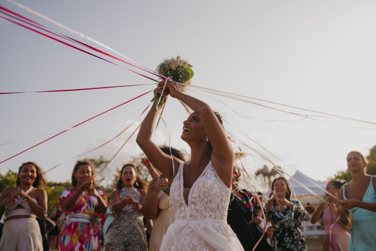A bride lifting her bouquet from a spread of coloured ribbons as guests look on, Myl'Evasions