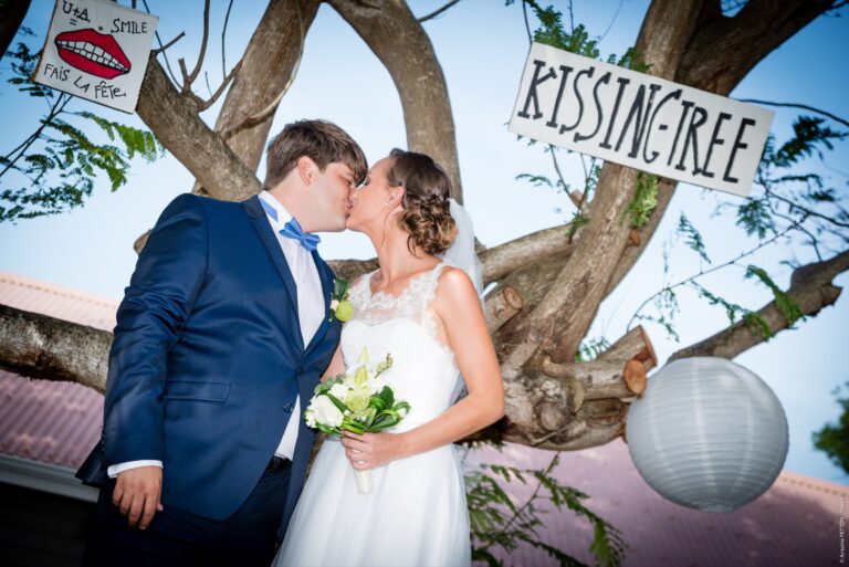 A couple kissing beneath a tree hung with a 'Kissing Tree' sign and paper lanterns, Myl'Evasions