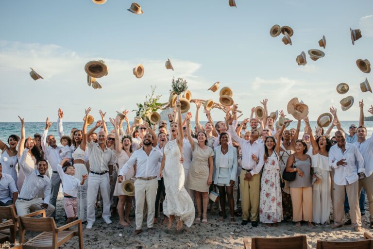 Guests in white throwing straw hats into the air on a beach, Myl'Evasions