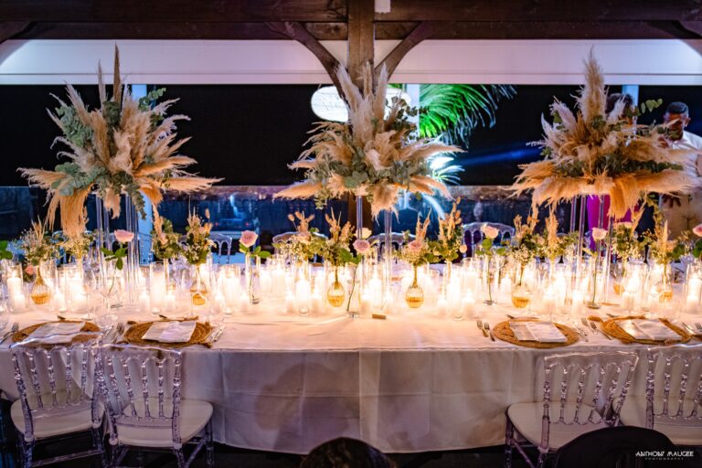 A candlelit banquet table dressed with pampas grass and tall floral arrangements under a timber roof, Myl'Evasions