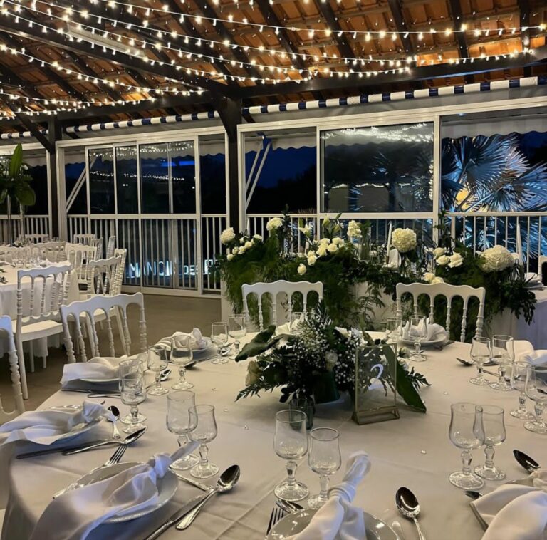 Round tables with white linen and greenery centrepieces in a glass-walled pavilion, Myl'Evasions