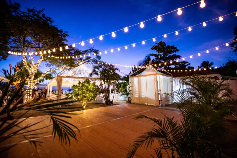 Festoon lights above a dance floor and white marquees at dusk in a tropical garden, Myl'Evasions