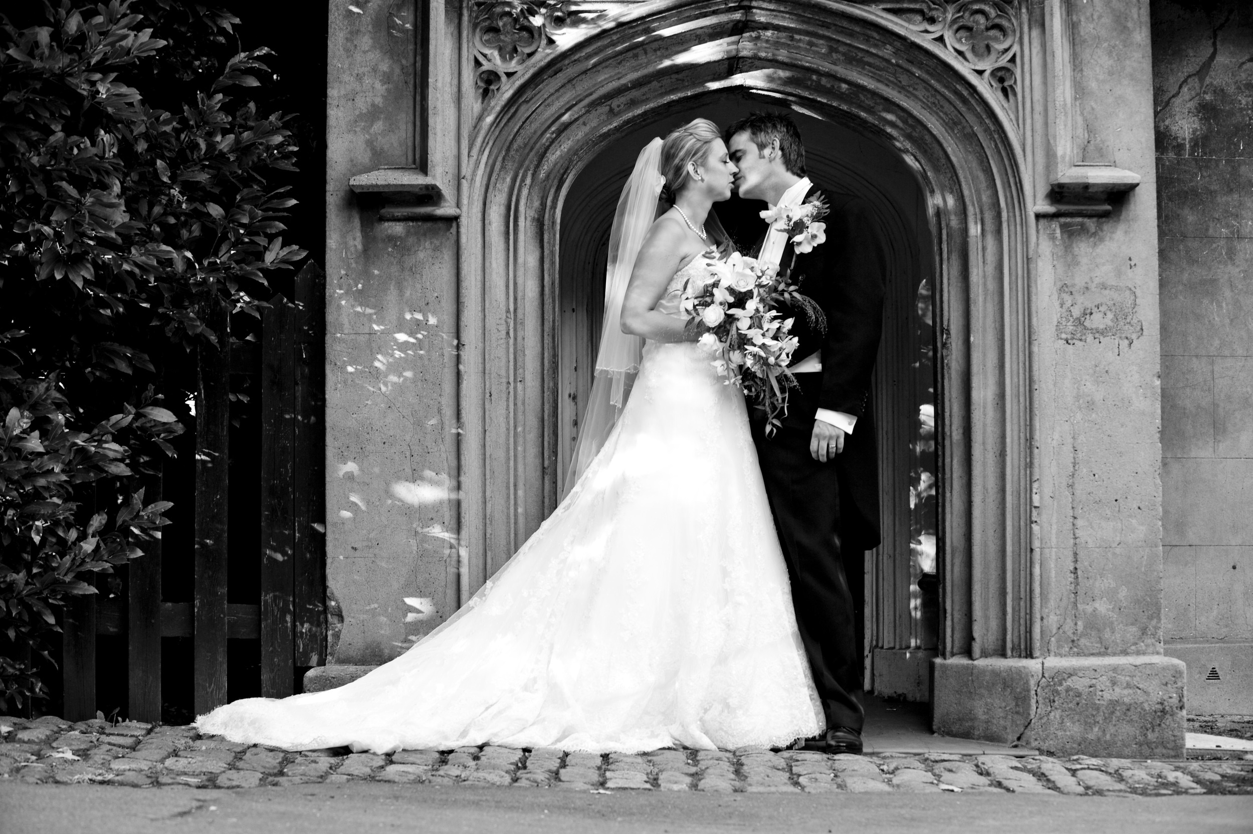 A bride and groom kissing beneath a stone archway, black and white, Nadine Pyke Make Up Artist