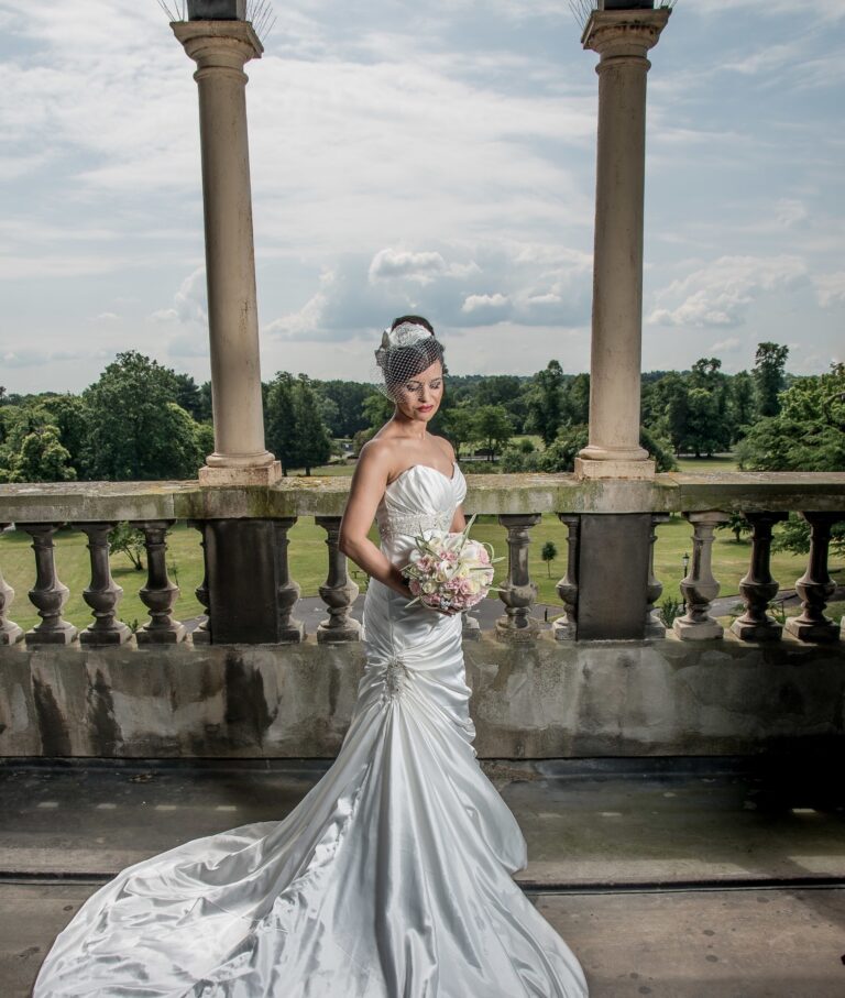 A bride in a fitted silver gown, holding her bouquet on a stone balcony, Nadine Pyke Make Up Artist