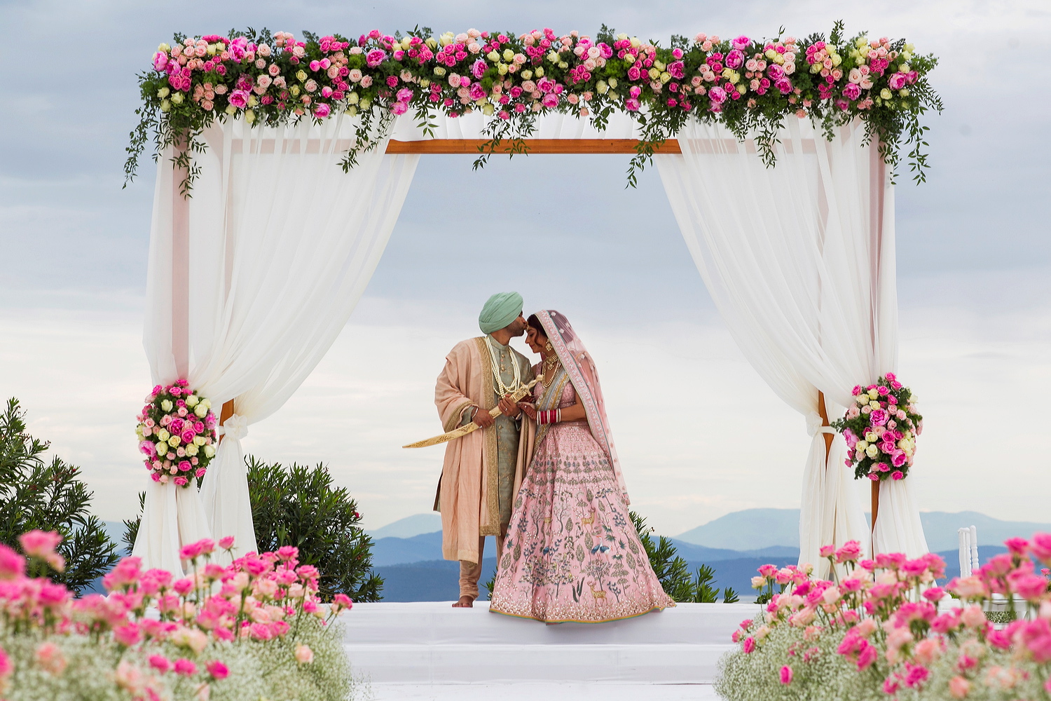 Couple beneath a floral mandap with pink flowers at the aisle edge, sea and hills behind, Nilyum Wedding and Event Design