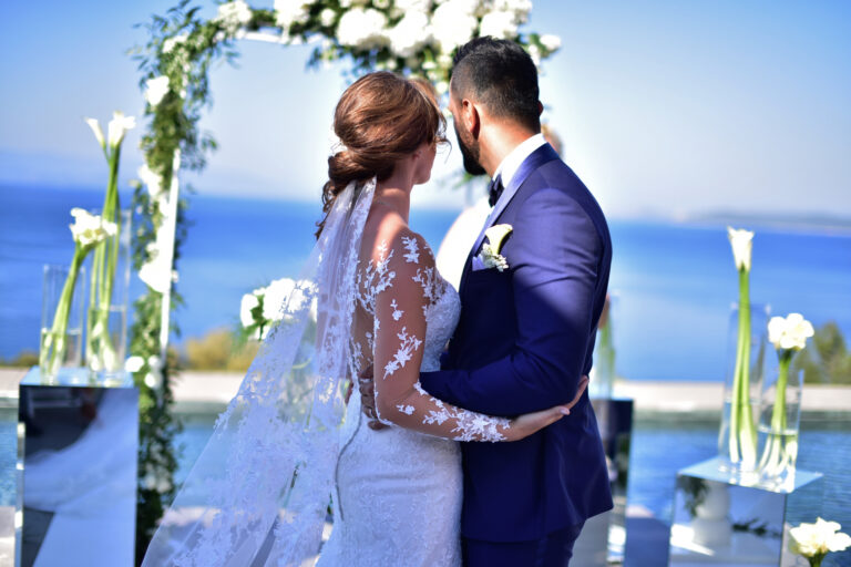 Bride and groom embracing in front of a floral arch with the sea behind, Nilyum Wedding and Event Design