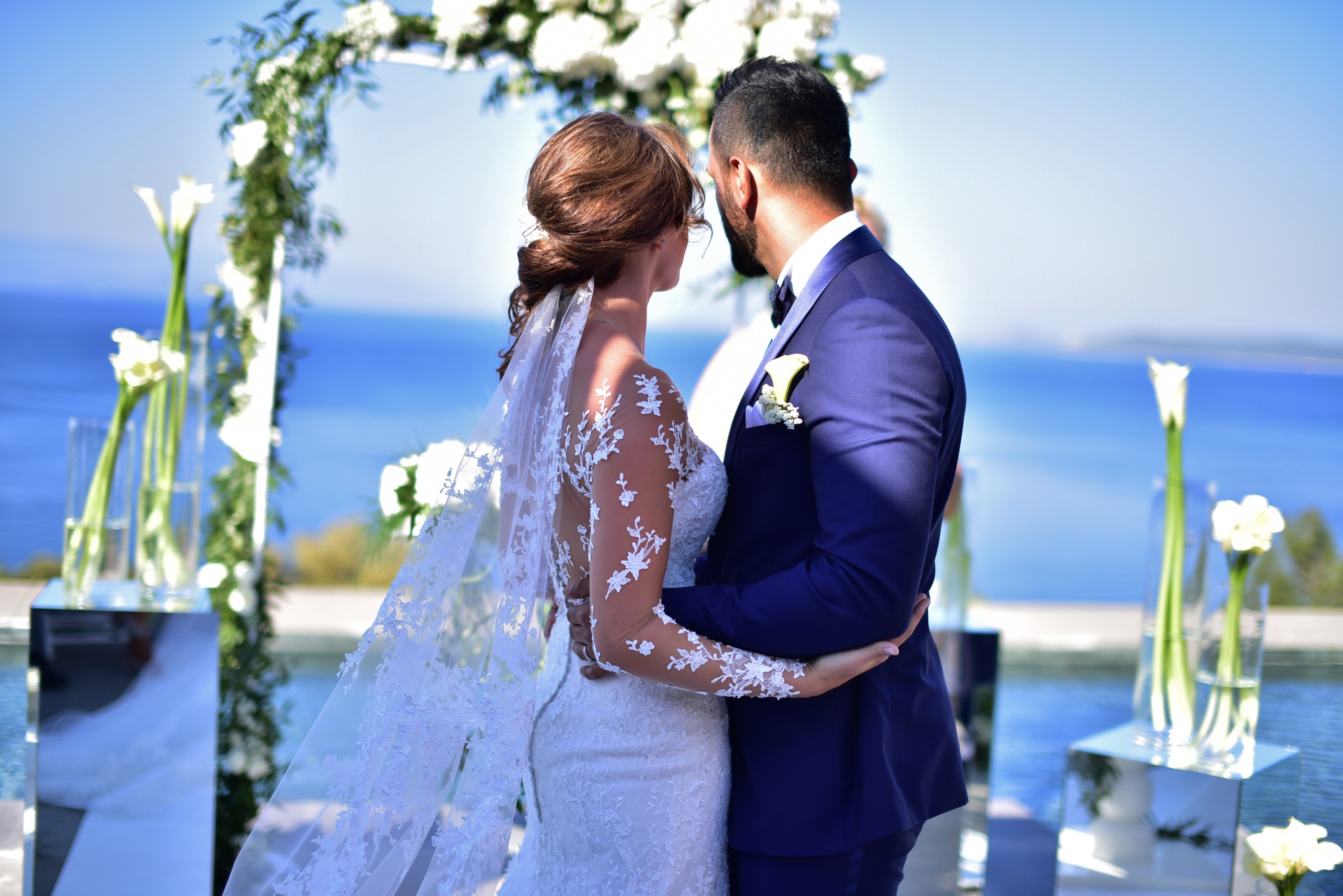 Bride and groom embracing in front of a floral arch with the sea behind, Nilyum Wedding and Event Design