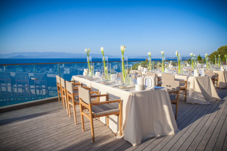 Long dining tables set along a sea-view terrace with tall glass vases and calla lilies, Nilyum Wedding and Event Design