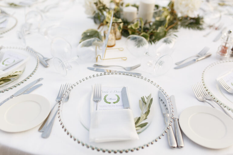 Round table set with white plates, silver chargers and olive sprigs, Nilyum Wedding and Event Design
