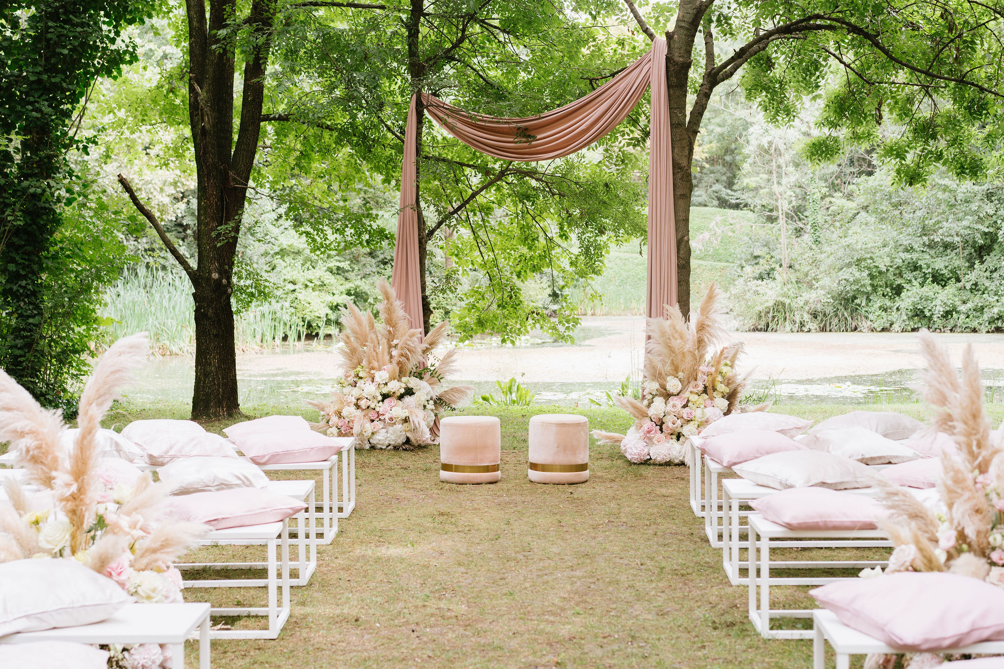A garden ceremony set-up: dusty pink drapery between two trees, pampas and roses, low white seats with cushions, a lily pond behind, Noemi Wedding