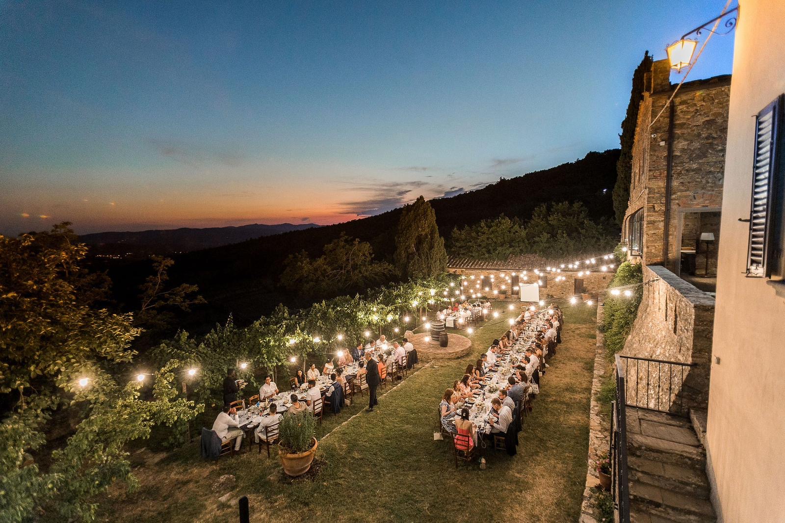 Long tables lit by festoon lights on the lawn below the house at dusk, hills fading behind, Montelucci Country Resort, Tuscany, Noemi Wedding