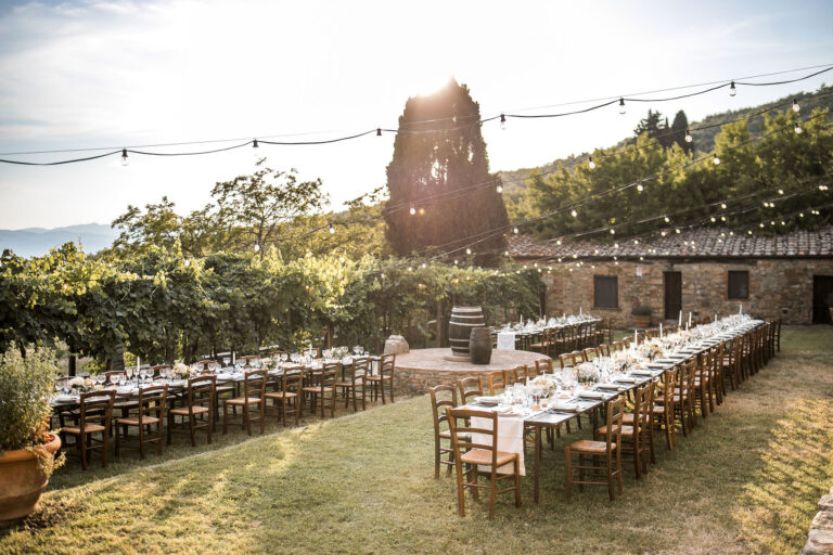 Long wooden tables on a lawn before a stone farmhouse, a wine barrel and festoon lights overhead, Noemi Wedding