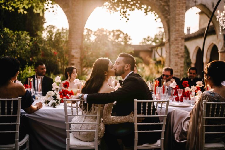 Dinner under a brick arcade at sunset, red roses and red candles on round tables, Noemi Wedding
