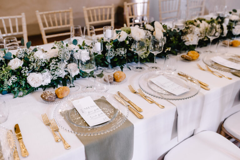 Place settings with glass chargers, gold cutlery and printed menus, white roses and greenery down the centre, Noemi Wedding