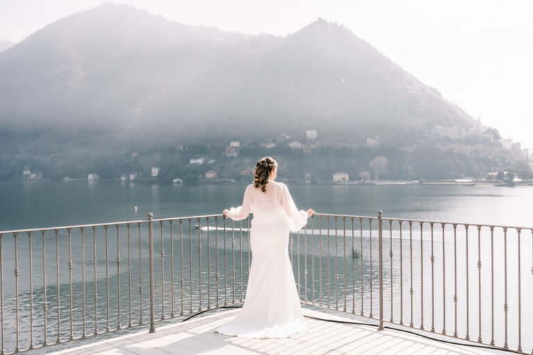 A bride alone at a lakeside railing, mist on the water and a wooded mountain opposite, Noemi Wedding