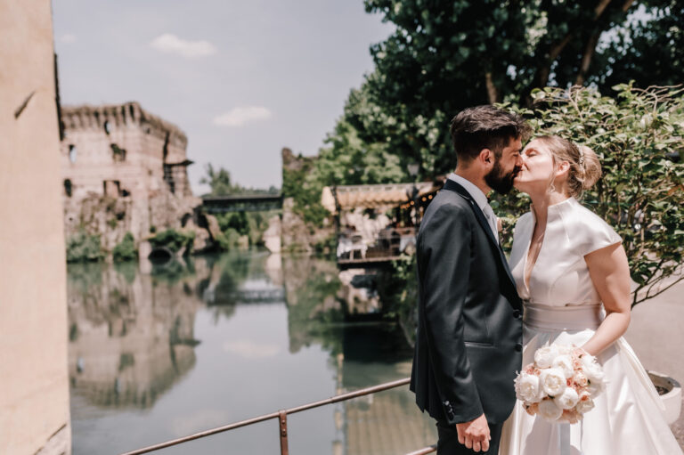 A couple by a river, an old stone tower and mill houses reflected in the water, Noemi Wedding