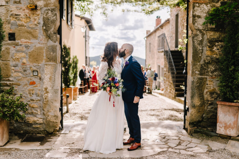 A kiss in a stone alley of a hill village, guests seated behind, Noemi Wedding