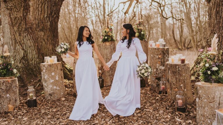 Two brides holding hands at a woodland ceremony set with candles on tree stumps, Nordic Adventure Weddings