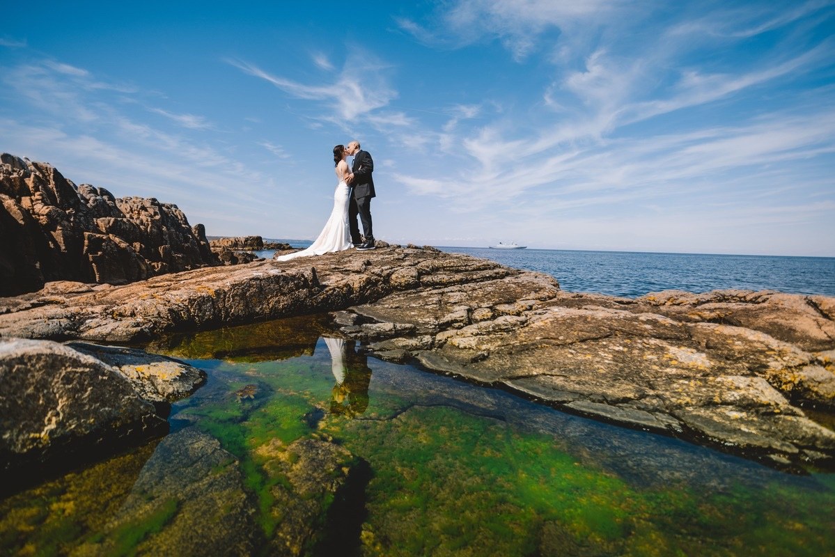 Couple embracing on a rocky shoreline beneath a blue sky, Nordic Adventure Weddings