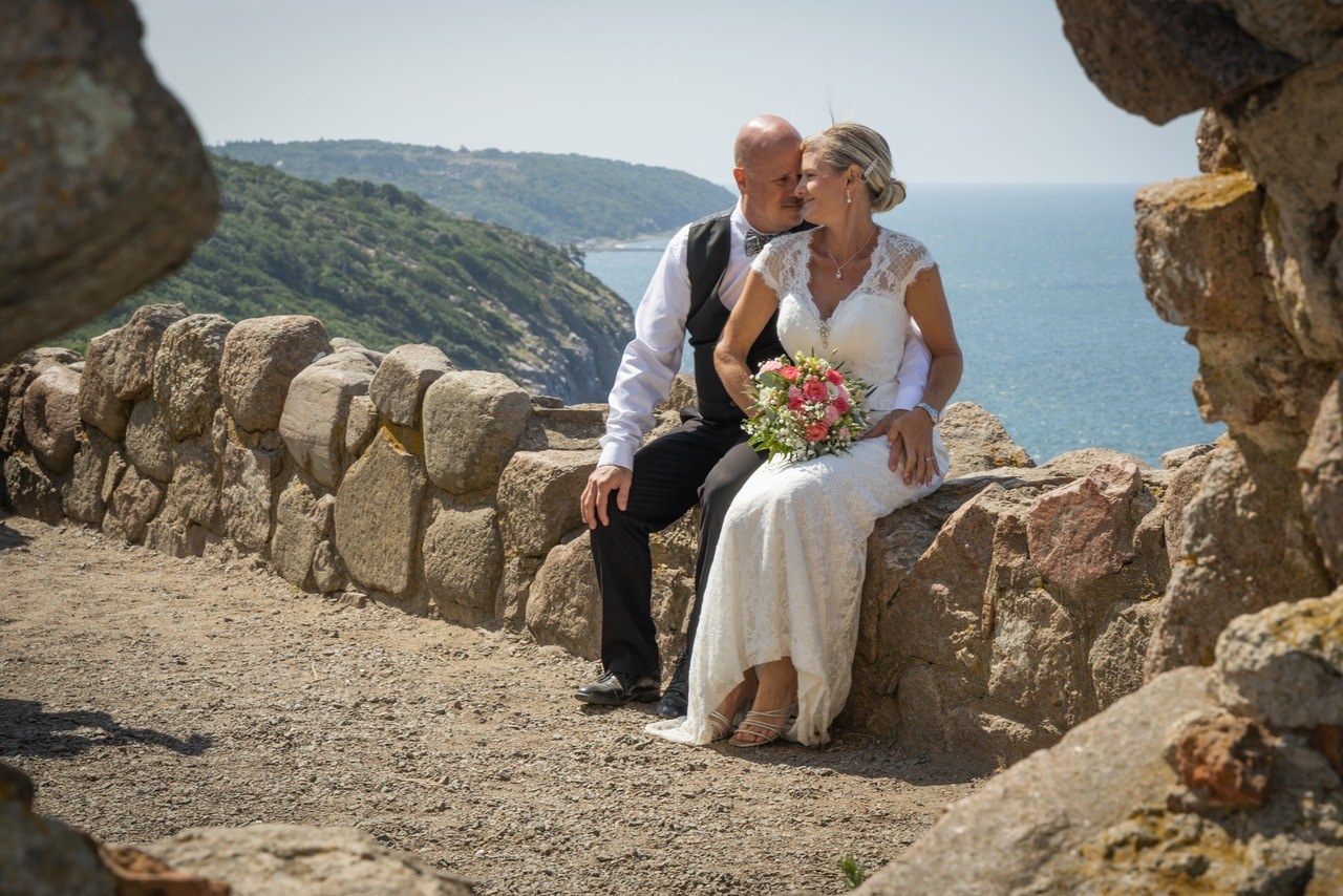 Couple seated on a stone wall above the sea, the bride holding a bouquet, Nordic Adventure Weddings