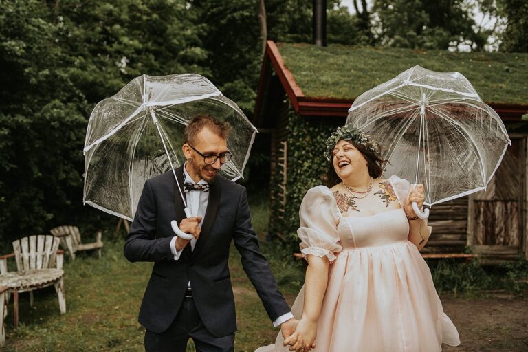 Couple laughing under clear umbrellas outside a turf-roofed cabin, Nordic Adventure Weddings