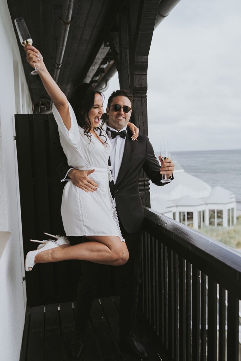 Couple celebrating with champagne on a balcony above the sea, Nordic Adventure Weddings