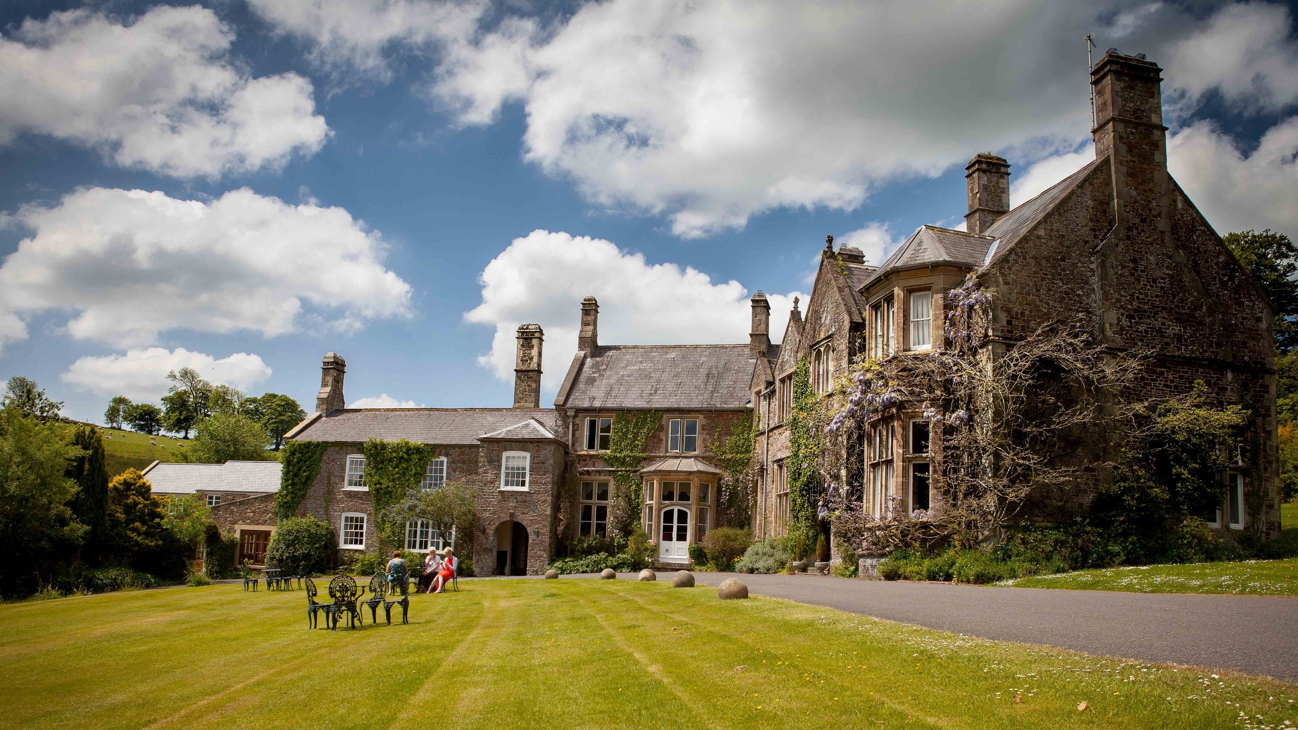 The ivy-clad manor house and its striped front lawn beside the gravel drive, with garden furniture set out on the grass, Northcote Manor Hotel