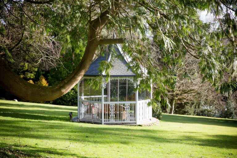 A white Victorian-style summerhouse on the lawn beneath the spreading branches of a large tree, Northcote Manor Hotel