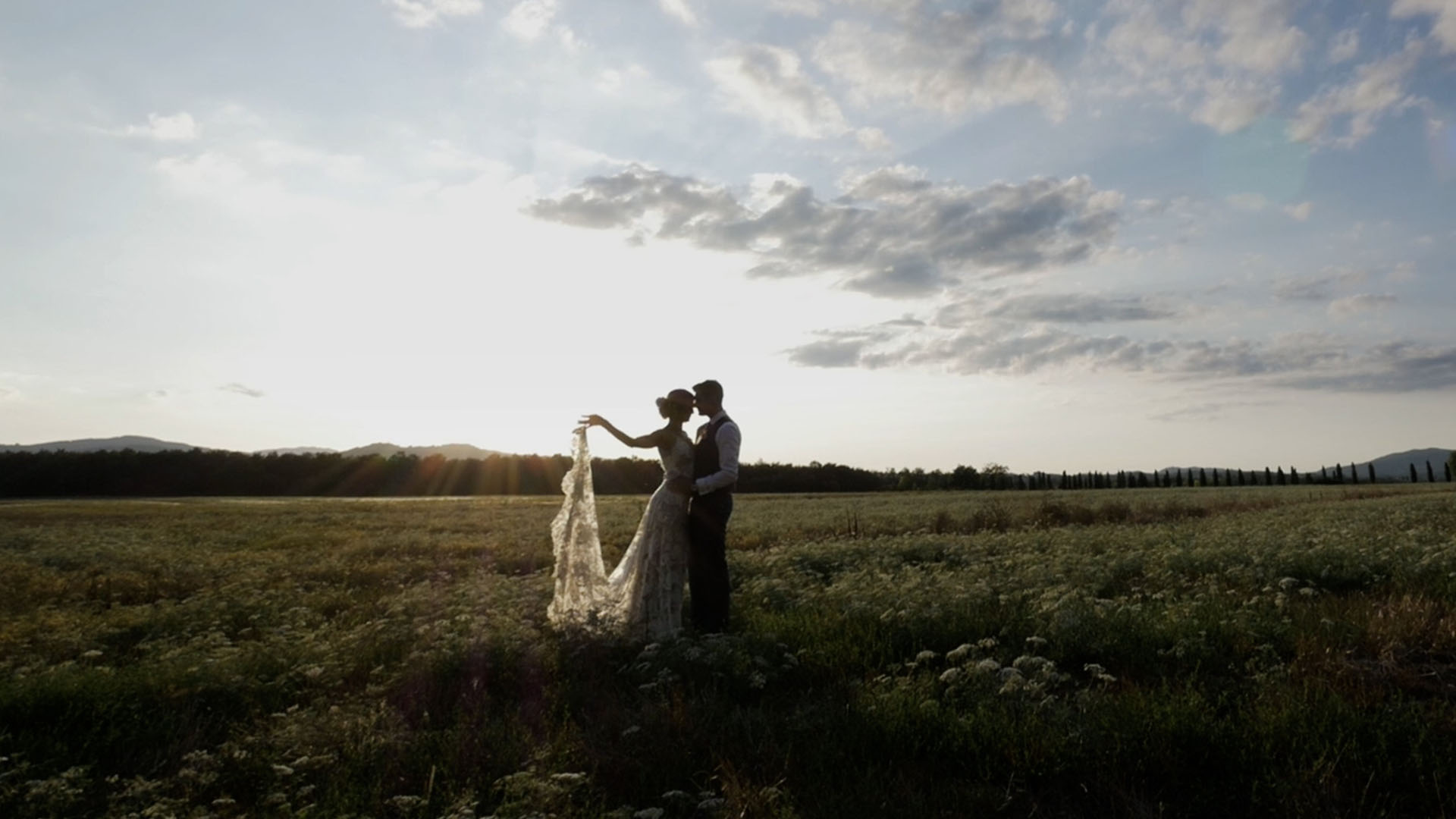 A couple in an open field at dusk, the bride's veil lifted by the wind, from a Nozze da Film wedding film, Nozze da film
