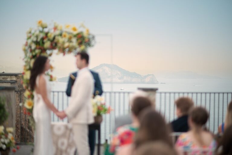 Hand in hand under a floral arch, a lighthouse cape across the water, O'Sarracino Events