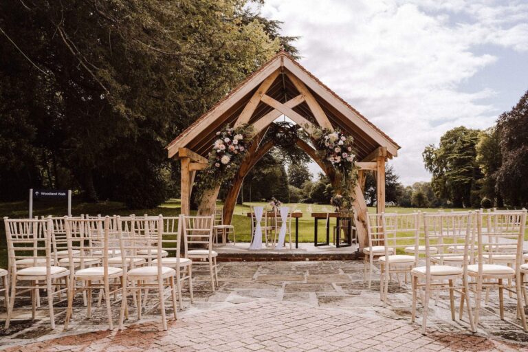 The oak-framed Woodland Pavilion dressed with flowers, with rows of white chairs on the paved ceremony area, Oakley Hall Hotel