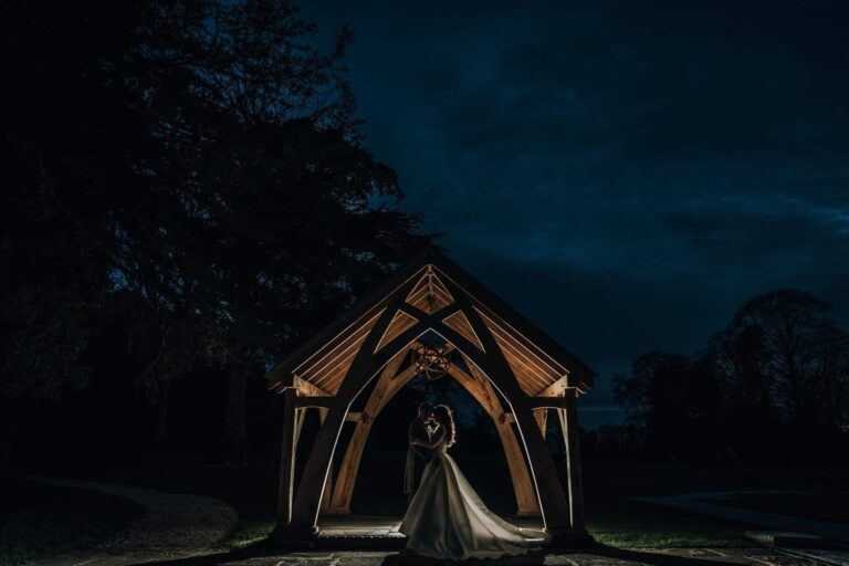 The Woodland Pavilion lit after dark, with a couple framed under the oak beams, Oakley Hall Hotel