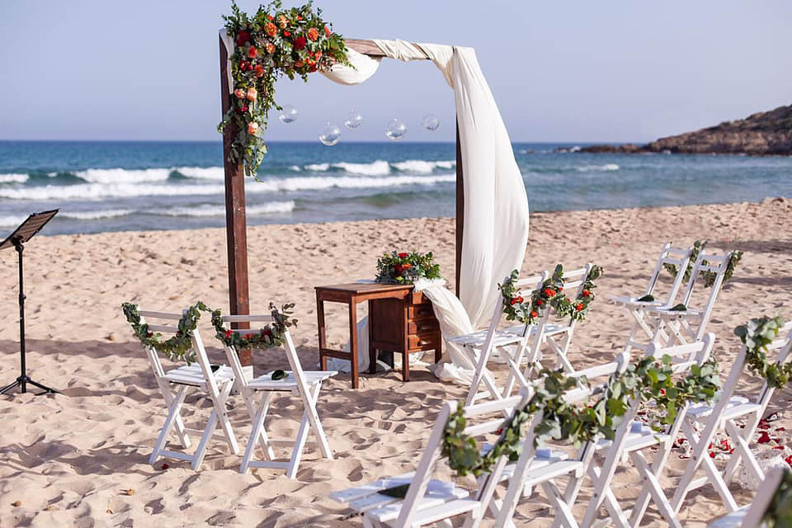 A beach ceremony set-up with a flowered wooden arch and white folding chairs, Sardinia, Oggi Sposi & Events