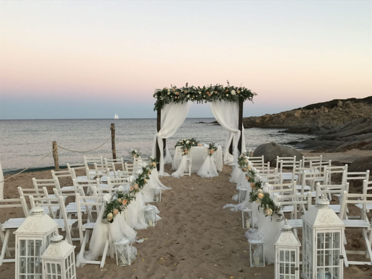 Beach aisle lined with white lanterns and greenery, leading to a flowered canopy at dusk, Oggi Sposi & Events