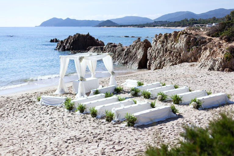 Ceremony benches and a white canopy set on a pebble beach among red rocks, Oggi Sposi & Events