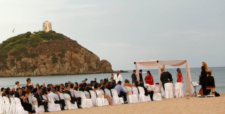 Guests seated for a beach ceremony below a coastal watchtower, Sardinia, Oggi Sposi & Events